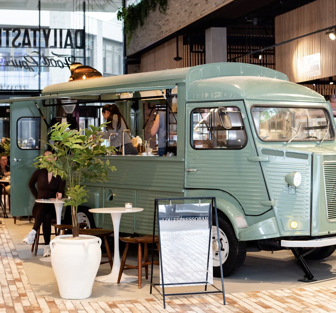 The sage green Citroën HY van that houses Loué Espressobar, serving through its open side window inside a market hall, with a sandwich board reading Lou.Espressobar out front